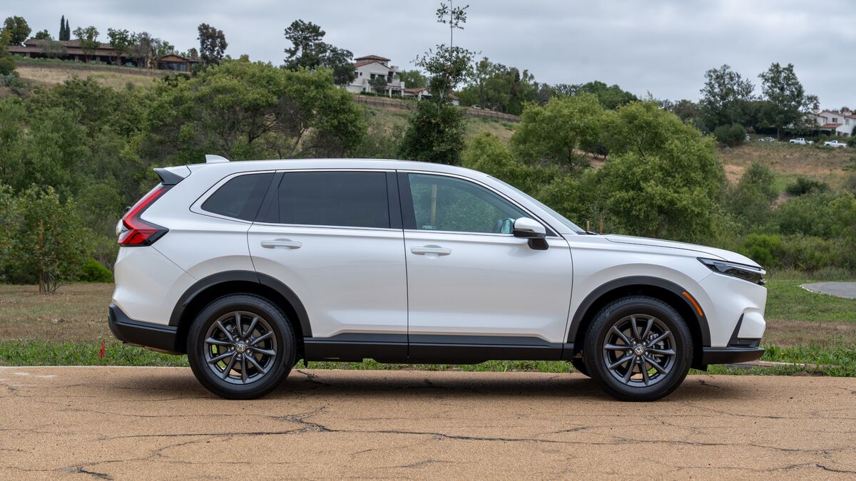 2026 Honda CR-V front right-side profile view in white with green hills in the background under cloudy skies.