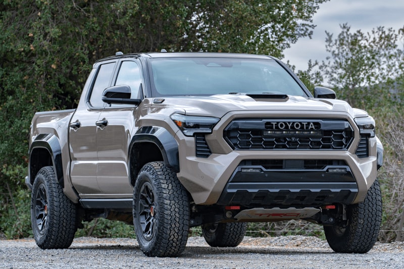 A front right-side 3/4 view of a 2026 Toyota Tacoma TRD Pro against a tree-filled backdrop under cloudy skies.