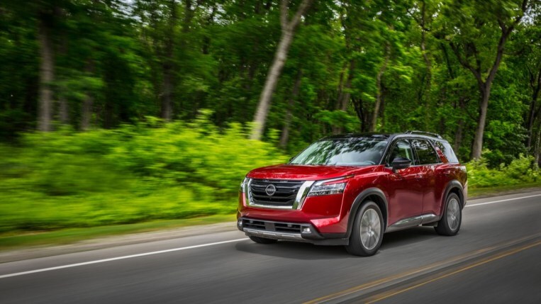 A red 2022 Nissan Pathfinder SUV drives through a forest. We see it from a three-quarter angle in front of the vehicle. It is boxy and rugged-looking with a large grille