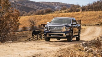A 2022 Nissan Titan pickup in dark blue passes some horses on a dirt road. The truck has a large chrome grille, chrome side mirror caps, and chrome running boards.