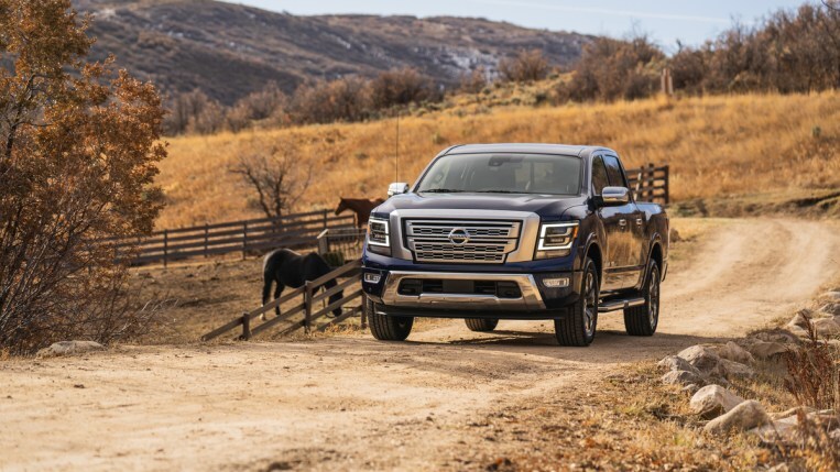 A 2022 Nissan Titan pickup in dark blue passes some horses on a dirt road. The truck has a large chrome grille, chrome side mirror caps, and chrome running boards.