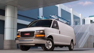 A GMC Savana van carrying two ladders sits parked in front of an office building. The van is plain white with no markings.