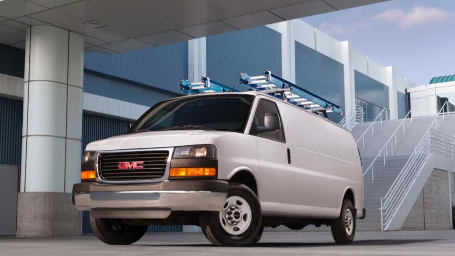 A GMC Savana van carrying two ladders sits parked in front of an office building. The van is plain white with no markings.