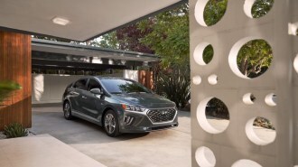 A 2018 Hyundai Ioniq hybrid sits parked in a driveway. The car is medium gray. We see it from a front quarter angle, facing slightly to our right. In the foreground, our view is partially blocked by an ornate decorative column.