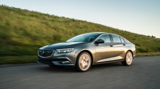 A 2019 Buick Regal drives past a grassy ridge. The car is medium gray. We see it in profile, facing to our left.