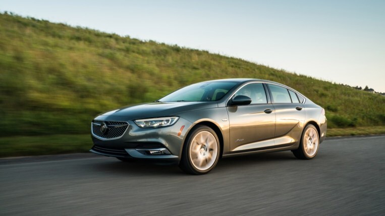 A 2019 Buick Regal drives past a grassy ridge. The car is medium gray. We see it in profile, facing to our left.