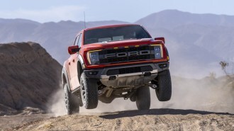 The 2021 Ford F-150 Raptor jumps on a sand dune. The truck is orange, with its front wheels nearly a foot off the ground. We see it from a nearly head-on angle.