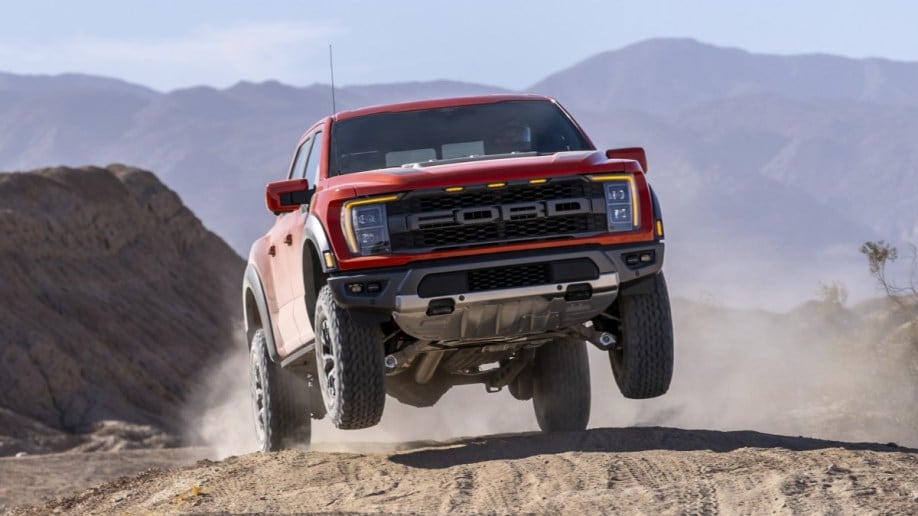The 2021 Ford F-150 Raptor jumps on a sand dune. The truck is orange, with its front wheels nearly a foot off the ground. We see it from a nearly head-on angle.