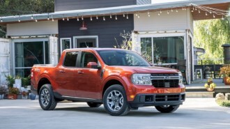 A 2022 Ford Maverick pickup sits parked in front of a house. The truck is bright orange. We see it from a front quarter angle, facing slightly to our right.
