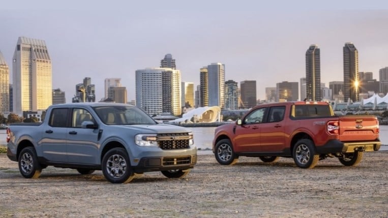 A pair of Ford Mavericks sit parked in front of a city skyline. On the right is an orange truck facing away from us so we see its left side. On the left is a powder-blue truck, positioned so that we see its right side.