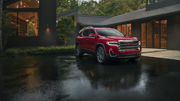 A 2022 GMC Acadia sits parked in the driveway of an elaborate modernist home. The SUV is red, with a large chrome grille. We see it from a front quarter angle, facing slightly to our right.