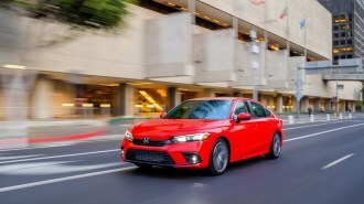 A 2022 Honda Civic drives on a city street. The car is bright red. We see it from a front quarter angle, facing slightly to our left.