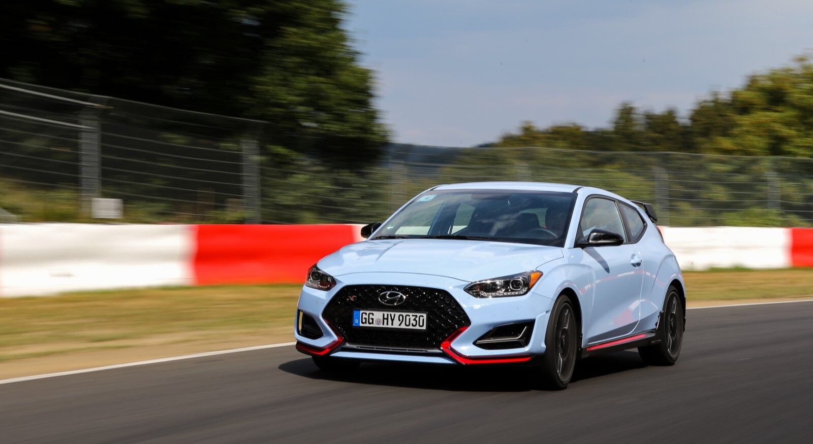 A Hyundai Veloster N performance hatchback races around a testing track. The car is powder blue with red accents low on its front fascia and side skirts.