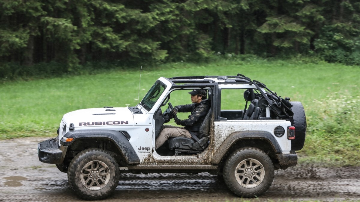 A two-door Jeep Wrangler drives through mud with its doors removed. The SUV is white. We see it in profile.