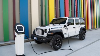 A 2022 Jeep Wrangler 4xe plug-in hybrid sits plugged in to charge. The Jeep is white and has four doors.