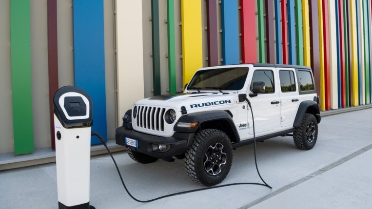 A 2022 Jeep Wrangler 4xe plug-in hybrid sits plugged in to charge. The Jeep is white and has four doors. 