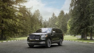 A 2021 Lincoln Navigator sits parked alone in a lot surrounded by forest. The big SUV is black with chrome highlights. We see it from a front quarter angle, facing to our left.