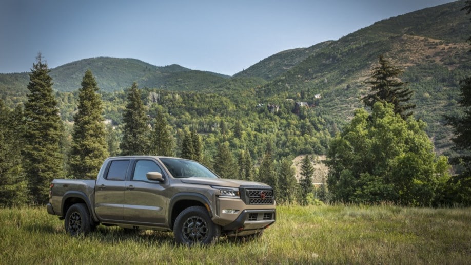 A 2022 Nissan Frontier pickup sits parked in a grassy field. The truck is a muted green color. We see it from a front quarter angle, facing to our right,