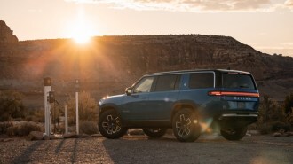 A Rivian R1S electric SUV sits plugged into a charger in a gravel parking lot against a canyon backdrop. The SUV is medium blue. We see it from a rear quarter angle.