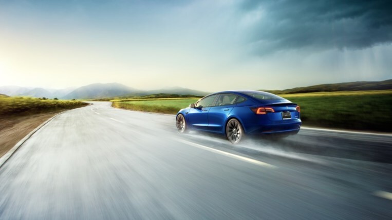 A Tesla Model 3 drives on a wet road. We see the car from a rear quarter angle, facing to our left and driving away from us. It is medium blue and kicking up water from its tires.