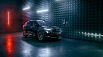 A Chevy Bolt EUV sits, plugged into a wall charger. The car is black and the lighting is dark. We see its right side in profile.
