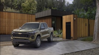 The 2023 Chevy Colorado sits parked in front of a modernist home. The truck is a muted green color, with a busy hood full of bulges and creases