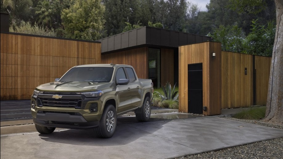 The 2023 Chevy Colorado sits parked in front of a modernist home. The truck is a muted green color, with a busy hood full of bulges and creases