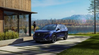 A 2023 Honda CR-V sits parked at a house on a lake. The car is blue. We see it from a front quarter angle, facing slightly to our left. A man is walking away from it, talking on a phone.