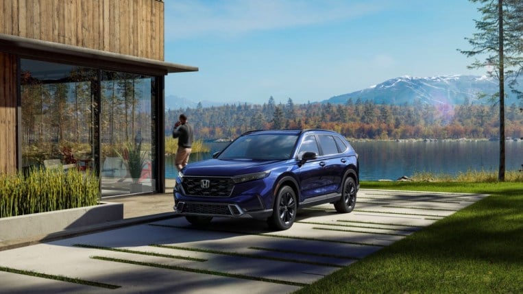 A 2023 Honda CR-V sits parked at a house on a lake. The car is blue. We see it from a front quarter angle, facing slightly to our left. A man is walking away from it, talking on a phone.