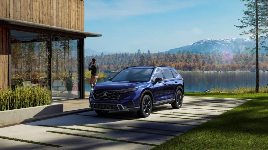 A 2023 Honda CR-V sits parked at a house on a lake. The car is blue. We see it from a front quarter angle, facing slightly to our left. A man is walking away from it, talking on a phone.