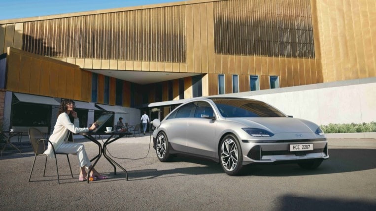 The 2023 Hyundai Ioniq 6 sits parked outside a modernist building next to a woman on a large tablet at a folding table. The car is silver. We see it from a front quarter angle, facing slightly to our right. It is extremely curvy and aerodynamic.