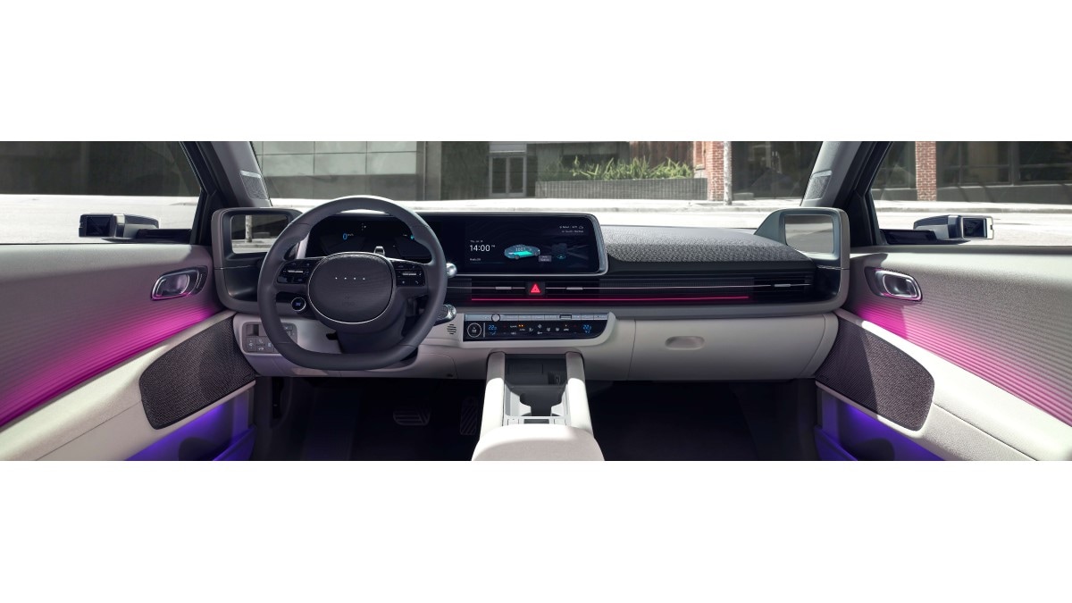 The interior of the 2023 Hyundai Ioniq 6. We see the dashboard from between the front seats. The cabin is upholstered in an off-white and light gray scheme, with purple ambient lighting.
