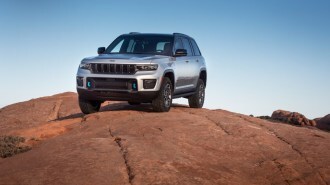 A 2023 Jeep Grand Cherokee 4xe Trailhawk sits parked on a rocky slope. The SUV faces slightly to our left. It's silver, with a black matte hood to reduce glare and bright blue tow hooks.