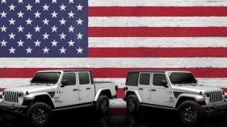 The 2023 Jeep Wrangler and Jeep Gladiator Freedom Editions are parked against an American flag backdrop. Both are silver with small black and white American flag decals ahead of the doors. The SUV faces right, the truck left.