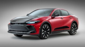 The 2023 Toyota Crown sits parked in front of a gray backdop. The car is shaped like a sedan but has an SUV's high ground clearance. It is red with a black hood and front fascia. We see it from a front quarter angle, facing slightly to our left.