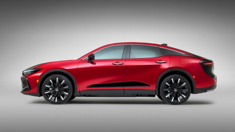 The 2023 Toyota Crown sits parked in front of a gray backdop. It sits high like an SUV, though it is shaped like a sedan. It is red, with an angular black strake along the bottom of the doors.