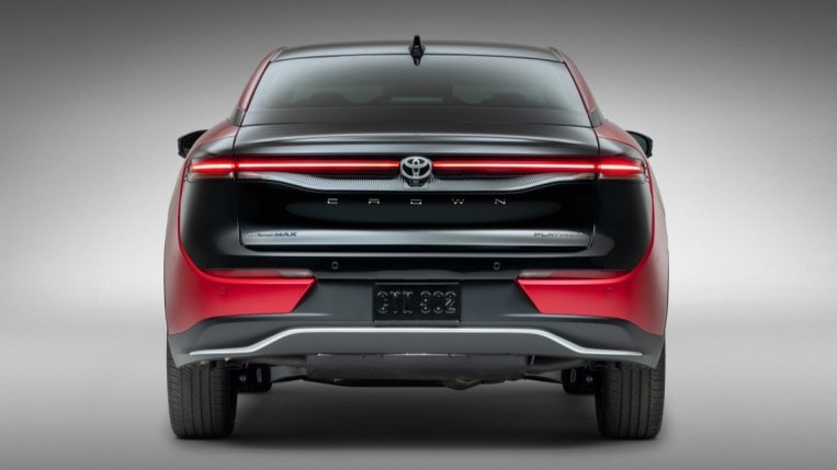The 2023 Toyota Crown sits parked in front of a gray backdop. We see it from the rear. The trunk lid is painted black, but the sides of the car are red.