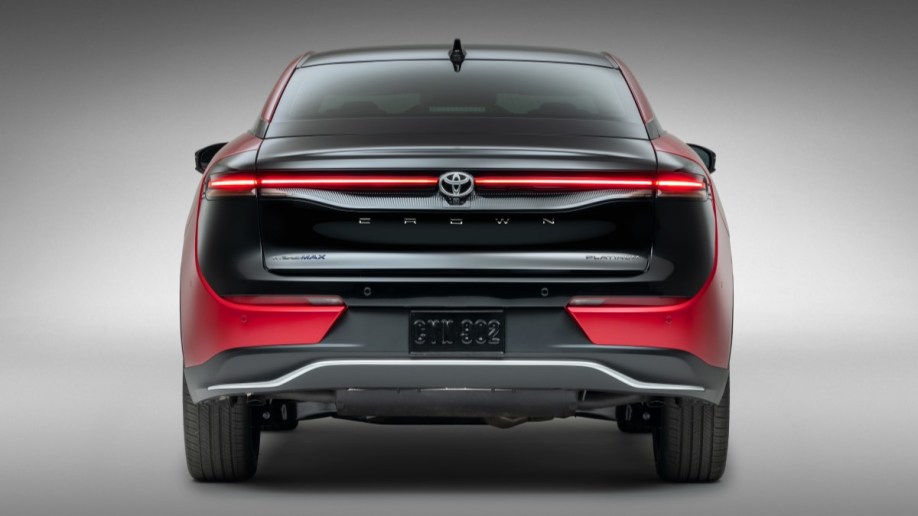 The 2023 Toyota Crown sits parked in front of a gray backdop. We see it from the rear. The trunk lid is painted black, but the sides of the car are red.