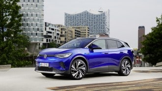 A 2023 VW ID.4 sits parked in front of a city skyline. The car is medium blue with a black roof. We see it in profile, facing to our left.