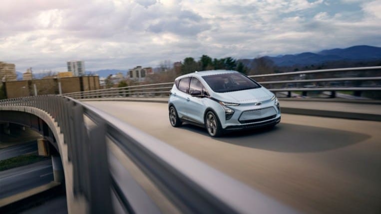 A Chevy Bolt EV drives away from a city on an overpass. The car is a very light blue. We see the car in focus and highway guadrails blurred to show motion.