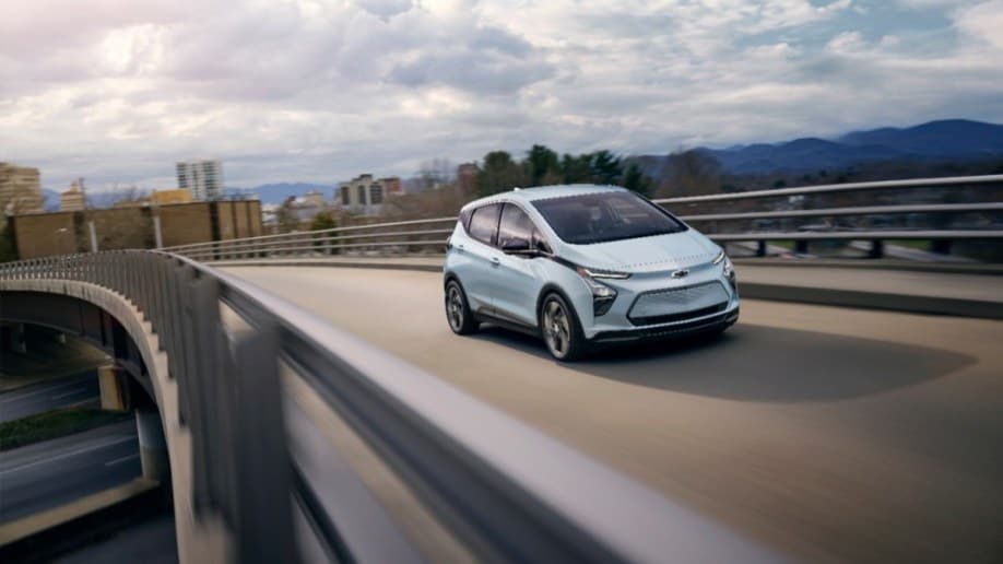 A Chevy Bolt EV drives away from a city on an overpass. The car is a very light blue. We see the car in focus and highway guadrails blurred to show motion.