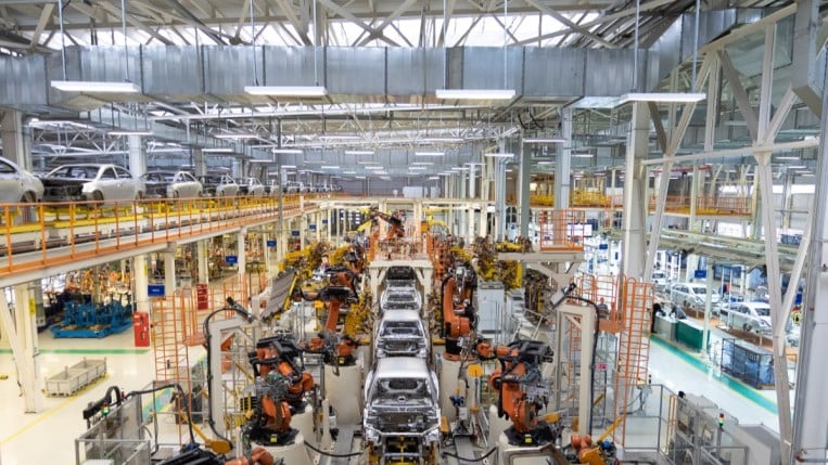 An overhead photo of a production line inside a car factory. We see workers operating two lines of orange robots on either side of a line of silver sedans.