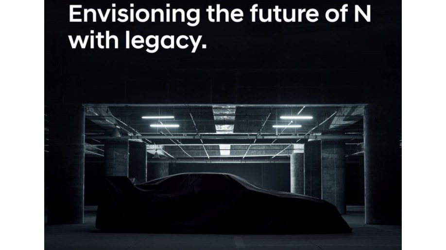A mystery car sits under a sheet in a parking garage. It seems to have the long, low lines of a sports coupe, and a high rear wing. Above it, text reads "Envisioning the future of N with Legacy"