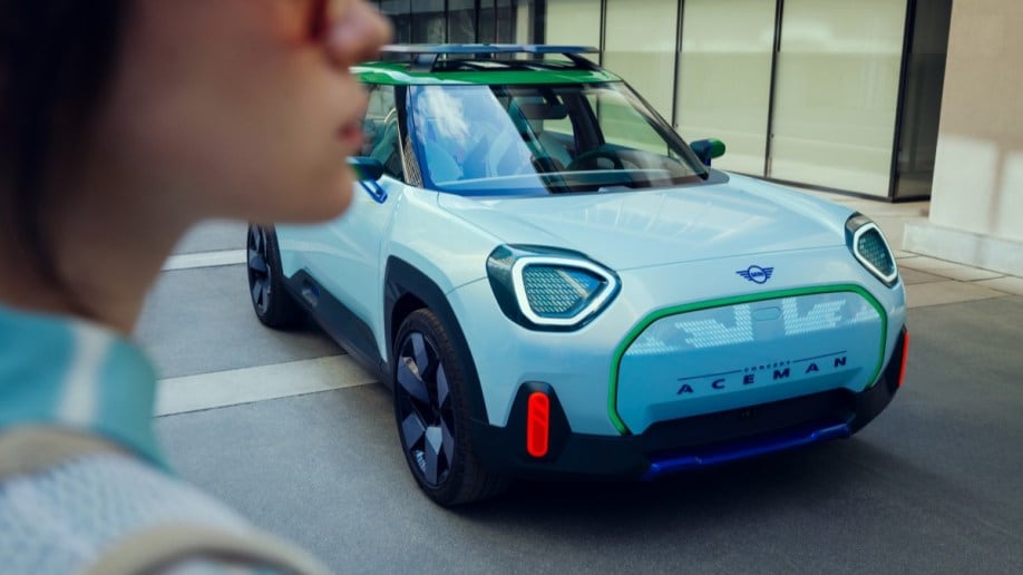 The Mini Aceman concept is parked next to a set of floor-to-cieling windows. We see its false grille, made of LED lights in the shape of the Union Jack. A woman is walking past in the foreground, slightly out of focus.