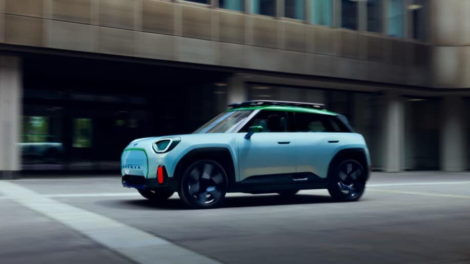 The Mini Aceman concept drives past a brutalist office building. The car is white with a green roof, in the classic Mini shape but with the rear pillars canted forward slightly.