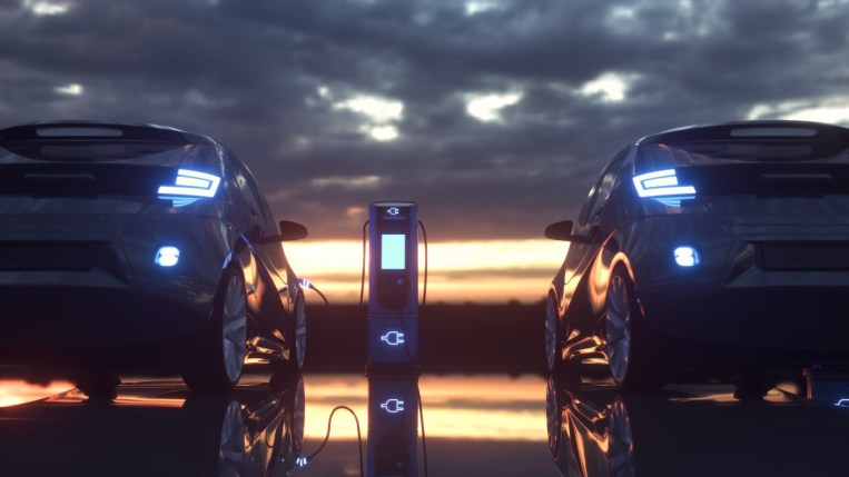 A pair of electric vehicles sit parked against a sunset. We see them from the rear. The one on the left is plugged into a charging station.