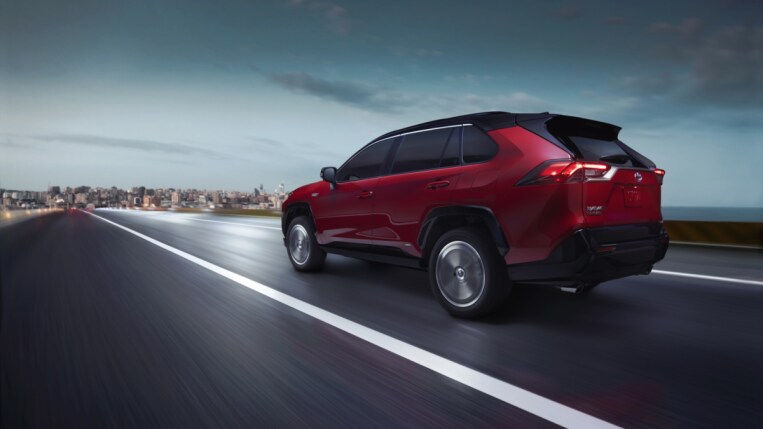 The 2022 Toyota RAV4 drives on a wet road. The SUV is read with a black roof. We see it from a rear quarter angle, driving away from us.