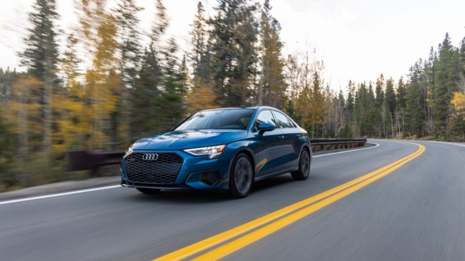 The 2022 Audi A3 sedan drives through a forest of evergreens. The car is a medium blue Audi calls "atoll blue." We see it from a front quarter angle, facing slightly to our left.