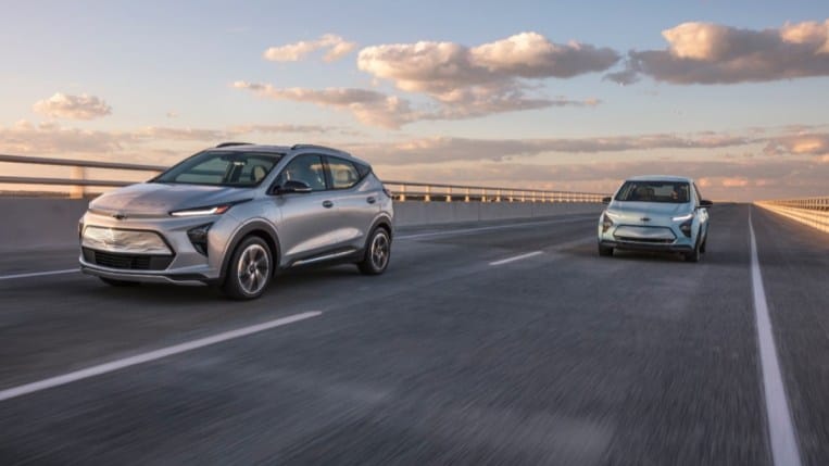 A 2022 Chevy Bolt EUV and a 2022 Chevy Bolt EV drive together on an elevated highway. Both cars are silver, and driving toward the camera at a slight angle facing to our left