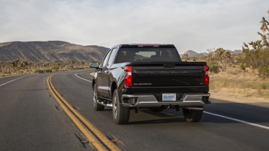 A 2022 Chevrolet Silverado pickup drives down a stretch of desert highway. The truck is black. We see it from a rear quarter angle as it drives away from us.